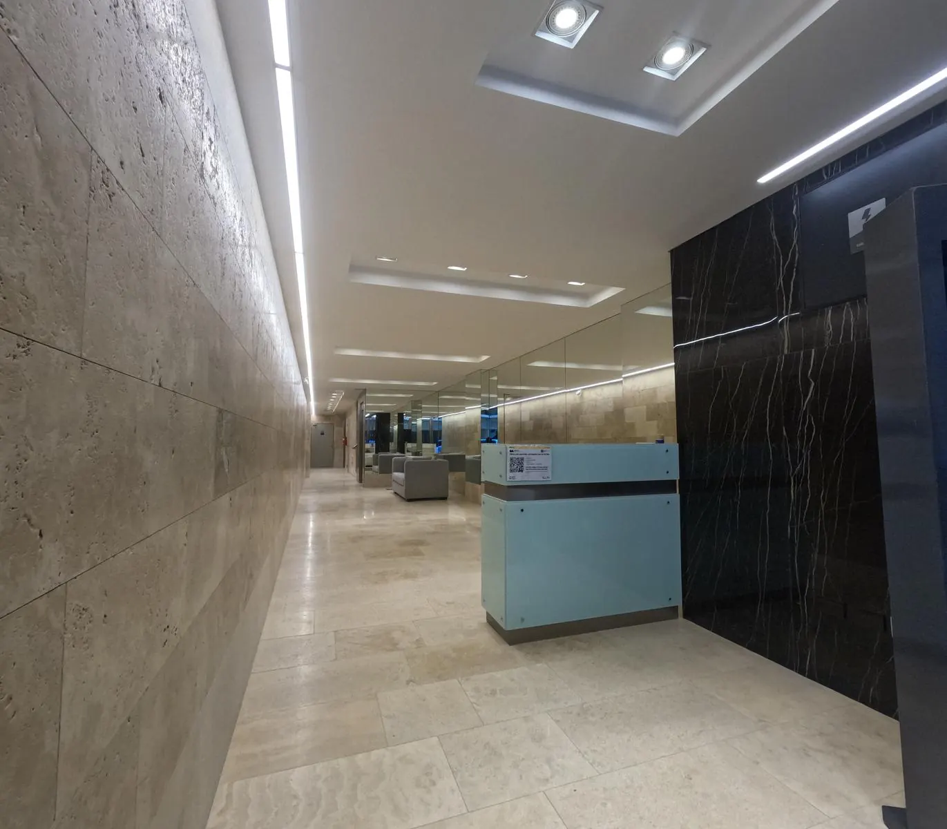 Hall de acceso terminado con revestimiento de travertino, mármol negro e iluminación lineal